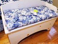 Front view of white wooden storage bench with blue and white floral tufted cushion on top.