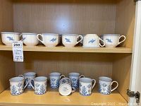 Shelf displaying 12 Villeroy & Boch Casa Azul tea cups on top shelf and 8 coffee mugs on bottom shelf with matching blue and white pattern showing detail clearly.