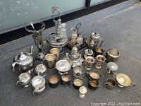 Wide view of entire lot showing all silver plate pieces including vases, teapots, cups, condiment holders, and trays on dark asphalt surface.