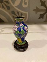 Front view of the blue cloisonné vase showing detailed pink and white floral designs around the body, gold-tone rim and base, on a round wooden stand.