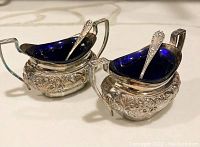Pair of silver salt cellars with cobalt blue glass liners and silver plated spoons inside, showing detailed floral engravings and design.