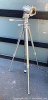 Full view of the aluminum tripod floor lamp standing outdoors on pavement with a metal wall background.