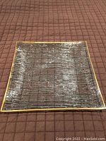 Square glass serving plate laid flat on a brown textured cloth background, showing the full top surface and square shape with gold trim edges.