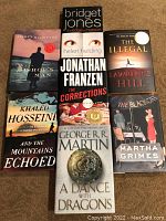 Photo showing the front covers of all 7 hardcover fiction books arranged in a grid on a carpeted floor.