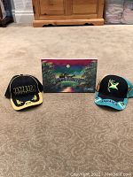 Front view of two Jamaica baseball caps and the signed canvas painting arranged on carpet.