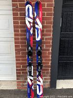 Pair of K2 Coomba skis showing top graphics red, blue, white, with bindings mounted, leaning against brick wall.