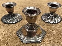 Set with one hexagonal sterling silver candlestick and two silver plated candlesticks with fan design bases on carpeted surface.
