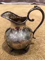 Front and side view of the silver plated creamer showing detailed engraved design and ornate handle.