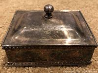 Front and angled view of the rectangular silver plated crumb collector with engraved lion rampant on lid and dentil decoration on sides.