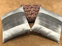 Two silver pleated throw pillows and one brown paisley accent pillow arranged on carpet.