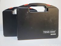 Two black TENS 3000 device carrying cases, stacked with one slightly behind the other. Cases have integrated handles and red locking tabs.