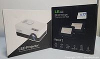 Photo showing white mini LED projector packaging and black packaging for LE Lighting Ever RGB LED Flood Lights, pack of 2.