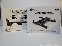 Photo showing two drone boxes: Holy Stone HS160 in black packaging and Idea 10 white drone packaging with visible branding and features listed.
