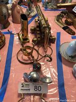 View of brass decorative items including a small cannon, candle holder, scorpion decor, and metal pieces on pink surface.