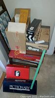 Photo of stacked board games and game boxes including Pictionary and Scrabble on chair and floor.