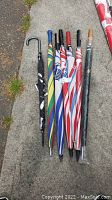 Seven umbrellas laid on concrete sidewalk: six golf umbrellas with straight handles and varying colorful canopy patterns and one smaller black umbrella with hooked handle.