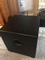 Black subwoofer set on carpeted floor with front-facing bass port, showing size and design.