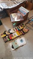 Photo showing a cardboard box filled with vinyl records along with a spread of 8-track tapes on the floor