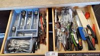 Overview of two adjacent kitchen drawers filled with assorted silverware and kitchen utensils, including knives, forks, spoons, peelers, and can opener.