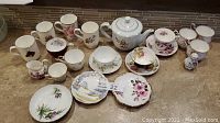 Photo showing assortment of porcelain teapot, teacups, saucers, and plates with various floral designs, arranged on a counter.