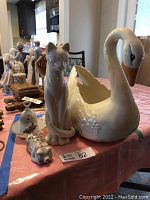 Photo showing large white porcelain swan figurine, sitting cat figurine, small pig figurine with floral designs, and white porcelain bell and bowl on a table.