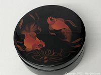 Top view of round black lacquer container lid with two gold fish and aquatic plant design in gold and brown tones, showing decorative details.