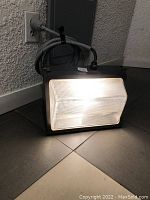 One outdoor wall pack LED light illuminated on a tiled floor next to a wall outlet with attached power cord.