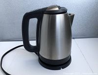Electric kettle with stainless steel body and black plastic handle and base shown from side view with power cord and plug visible.