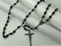 Overall view of the rosary laid on a white rolled fabric background showing black beads, chain links, centerpiece, and crucifix.