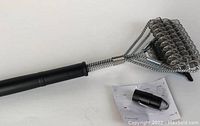 Photo showing the BBQ grill brush with black handle, twisted wire bristle head, additional plastic component, and instruction sheet on white background.