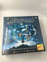 Front of Dreams board game box showing title and artwork of a person in front of an open book with magical light visual effects on a night sky background.