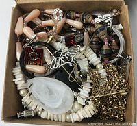 Small cardboard box with assorted jewelry including beaded necklaces, chains, brooches, and decorative pieces in various materials and colors.