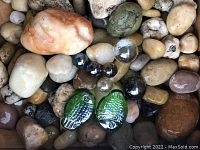 Assorted natural stones and marble balls with green glass fish among them, showing size and color variety.
