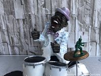 Front view of the figurine showing facial expression and upper body with drumsticks, part of the drum set