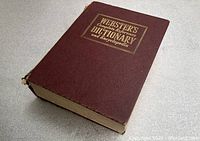 Angled view of closed Webster's dictionary book showing front cover with gold title and red textured cover.
