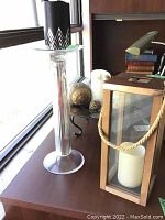 Tall clear glass candleholder with black candle and geometric pattern beside wooden lantern with pillar candle and table with decorative balls.