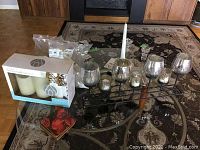 Photo showing metal candle holder rack with five small mercury glass votive holders, three large goblet-style glass candle holders, boxed three white pillar candles, two tall white taper candles, and some loose candles on a table.