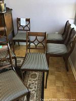 Six wooden dining chairs with upholstered seats. Chairs have a decorative backrest design with curved elements and striped upholstery. One chair shows damage requiring repair.