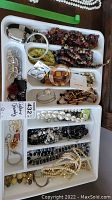 Photo shows two white trays filled with assorted costume jewelry including necklaces, bracelets, earrings, brooches, and a wristwatch. Items are arranged in plastic trays with multiple compartments.