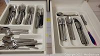 Two white plastic cutlery trays filled with forks, spoons, butter knives, and some knives with black and blue handles.