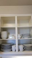 Shelves with neatly stacked white plates, bowls, and white mugs in a white cabinet.