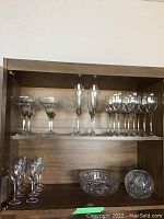 Full view of shelf with twenty-five glasses, including flutes, wine glasses, and liquor glasses, plus two crystal bowls on lower shelf