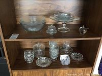 Wide view of wooden shelf holding glass and crystal bowls, containers, ash trays and decorative pieces including apples. Items are cleanable but currently dusty or slightly dirty.