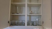 Glass cabinet shelves holding various clear drinking glasses including short tumblers and taller beer glasses.
