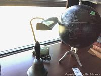 Photo showing the black globe, metal bell with wooden handle, and retro desk lamp on a surface near a window.