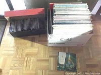 Photo showing a large white cardboard box filled with approximately 30 vinyl LP records standing upright with covers visible. Next to it is a smaller open box containing multiple CD jewel cases.