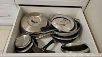 Drawer containing assorted stainless steel pots and pans stacked with lids and black handles visible