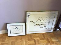 Pair of framed indigenous art prints resting on a hardwood floor against a beige wall. The smaller print measures approximately 11x14 inches, the larger about 20x24 inches. Both frames have beige mats and gold-colored frames.