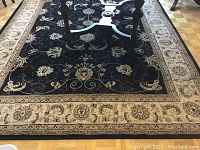 Photo of a heavy rectangular floral rug with a black base and cream border under a table.