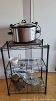 Full view of crock pot, warming plate, ceramic bowl and wire racks on a black metal shelving unit